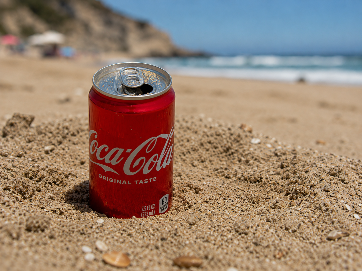 Red Coca-Cola can on a sandy beach with ocean and cliffs in the background, sand is on the top of the can